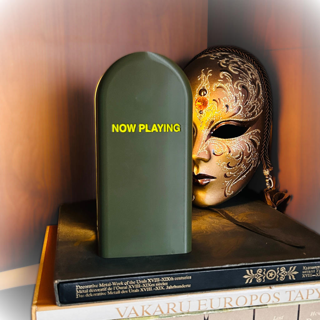 Decorative mask and green bookend with 'NOW PLAYING' text on a wooden surface.