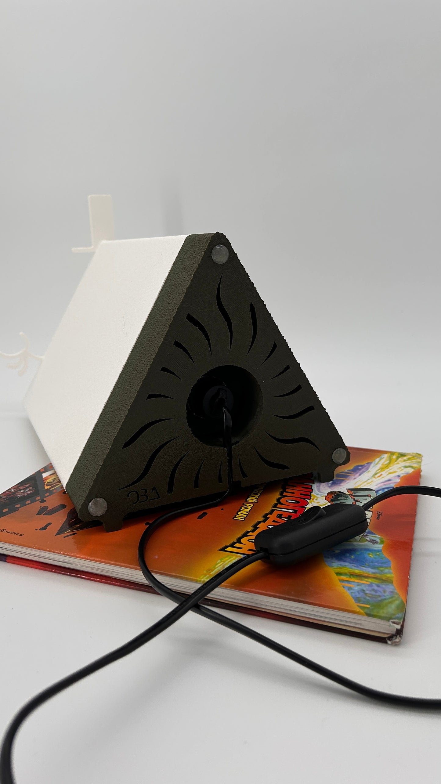 Triangular black device with a fan on top, connected to a cable, on a colorful book with a white background.