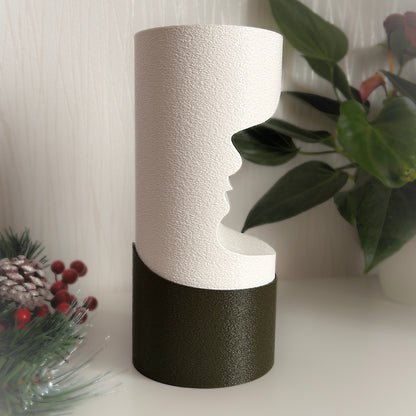 Decorative vase with textured white top and black base on a surface with plants and pinecones in the background.